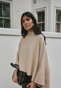 Poncho beige con un cuello alto, hecho de tela suave, presenta mangas anchas y se combina con un bolso negro. Fondo neutro.