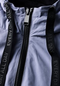 Ljuslila jacka med svart dragkedja och snoddar med texten "EVERYDAY ALL DAY". Slät textur med en elegant design.