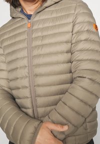 Donsjack in licht beige met een horizontaal doorgestikt ontwerp, ritssluiting met oranje accent en een capuchon. Zachte en gestructureerde stof.