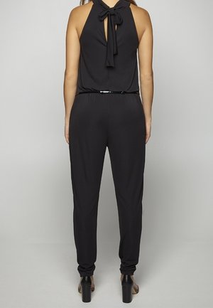 Femme debout, de dos, portant une combinaison noire sans manches avec un nœud noué au niveau du cou et des chaussures à talons hauts noires.