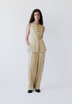 Sieviete ar cirtainiem matiem, tērpusies bēšā bezpiedurkņu pogātā topā un biksēs ar plašu kāju, stāv pret vienkāršu baltu fonu.