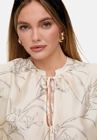 Cremefarbene Bluse mit schwarzen floralen Linienzeichnungen, einem Rundhalsausschnitt mit Binderverschluss und sanfter Drapierung. Sichtbare goldene Creolen-Ohrringe.