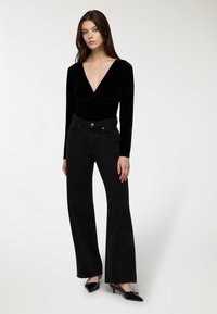 Młoda kobieta ubrana w czarną bluzkę z głębokim dekoltem w literę V i długim rękawem, czarne jeansy z wysokim stanem i szerokimi nogawkami oraz czarne szpilki z odkrytymi palcami.