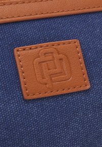 Sötétkék textil háttér, egy barna bőr címkével, rajta dombornyomott geometriai mintával. Az öltések körvonalazzák a címkét, ezzel textúrát adva neki.