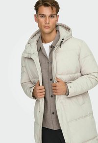 Lichtbeige gewatteerde jas met een capuchon, voorzien van een rits aan de voorkant, drukknopen en een gestructureerde binnenlaag. Soepele stof met een matte afwerking.