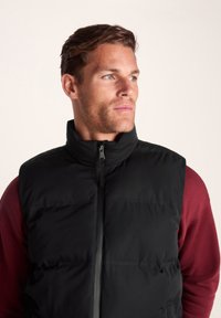 Black padded vest with high collar, front zipper, and side pockets. Worn over a dark red long-sleeve shirt, features a smooth texture.