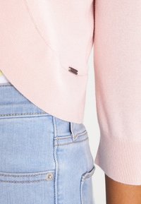 Ljusrosa stickad tröja med trekvartsärmar, som har en rundad nederkant och en liten metallflagga på vänster sida nära sömmen.