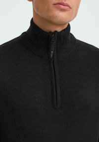 Black zip-up sweater with high collar, made of a textured knit fabric. Features a front zipper and minimal branding on the zipper pull.