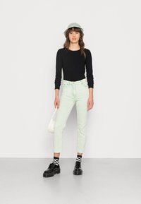 T-Shirt de manga longa preta com nervuras, calças de veludo cotelê verde claro, sapatos pretos com solas grossas e chapéu balde cinza. Bolsa branca simples como destaque.