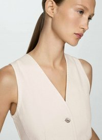 Robe sans manches couleur beige clair avec un décolleté en V profond et un bouton décoratif argenté. Présente une texture lisse et une silhouette ajustée.