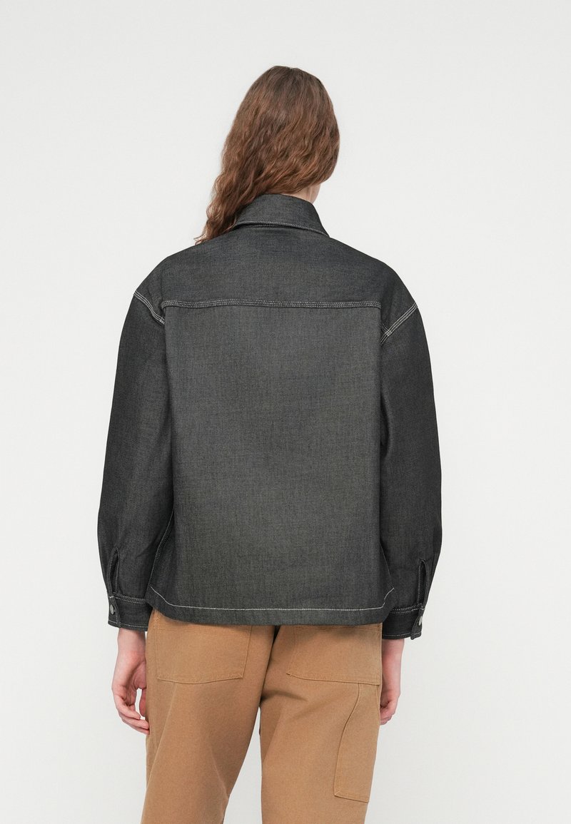 Sort denimjakke med løs pasform, synlige syningsdetaljer og knaplukning ved ærmerne, kombineret med lysebrune bukser. Bagfra ses krave og syninger.