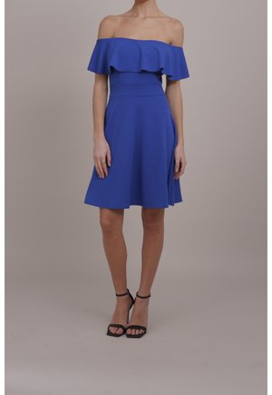 Vrouw draagt een koningsblauwe jurk tot op de knie, met een off-shoulder ontwerp en ruches, gecombineerd met zwarte hoge hakken met enkelriempjes.