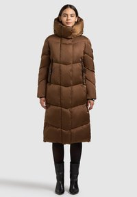 Rjava puhovka z visokim ovratnikom, geometrijskim prešitim vzorcem, žepki z zadrgo in dizajnom do kolen. Nošena s črnimi škornji.