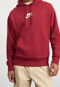 Röd bomullshuvtröja med framficka, justerbar dragsko i huvudet och vitt Nike-logotyp på bröstet. Texturerat tyg, avslappnad passform.