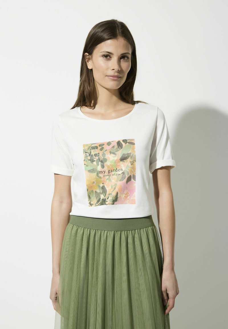 White T-shirt with short rolled sleeves, featuring a colorful floral print design and the text "my garden plenty of joy" in the center. Paired with a green, pleated skirt.