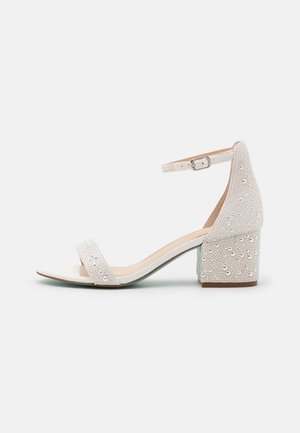 Bílé sandály na podpatku se širokým páskem a sponou na kotníku. Vyznačují se texturovaným svrškem s lesklými korálky a blokovým podpatkem.