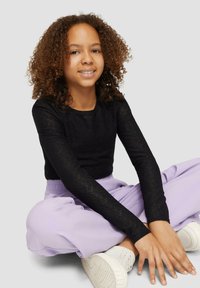 Zwarte longsleeve top met een gestructureerd ontwerp, gecombineerd met lichtpaarse oversized broek. Model draagt witte sneakers en zit met de handen op de knieën.