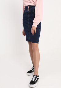 Mörk denim pennkjol med hög midja, prydd med en rad av tre guldknappar. Kombinerad med en ljusrosa tröja och svarta sneakers.