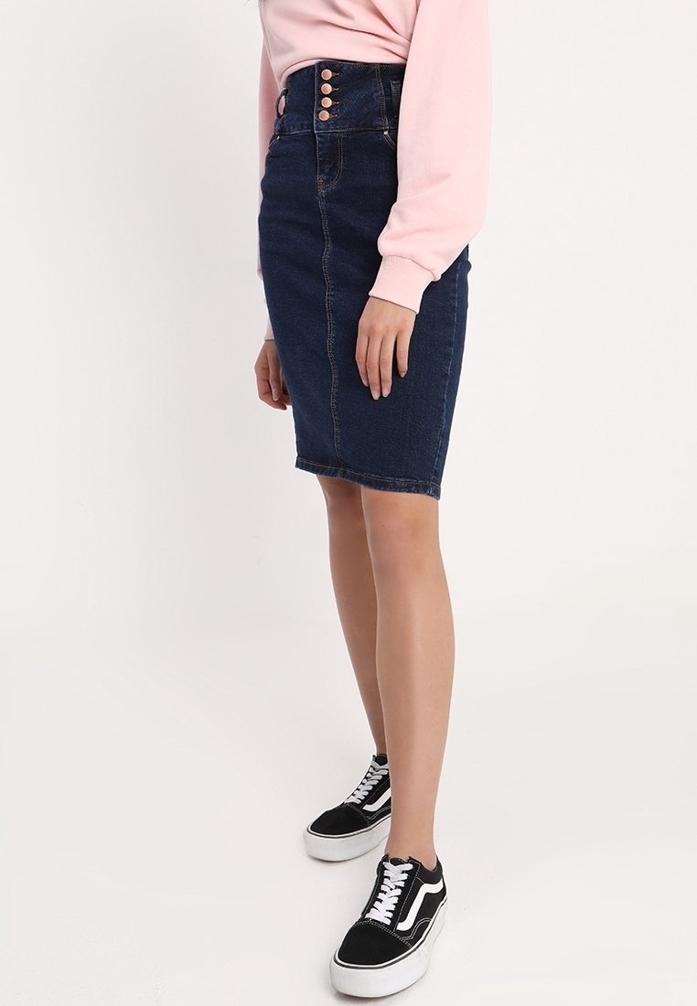 Mörk denim pennkjol med hög midja, prydd med en rad av tre guldknappar. Kombinerad med en ljusrosa tröja och svarta sneakers.