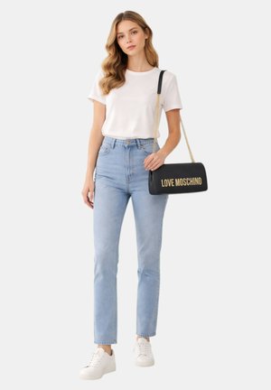 Giovane donna con t-shirt bianca, jeans azzurri, sneakers bianche, che tiene una borsa a tracolla nera Love Moschino con catena dorata.