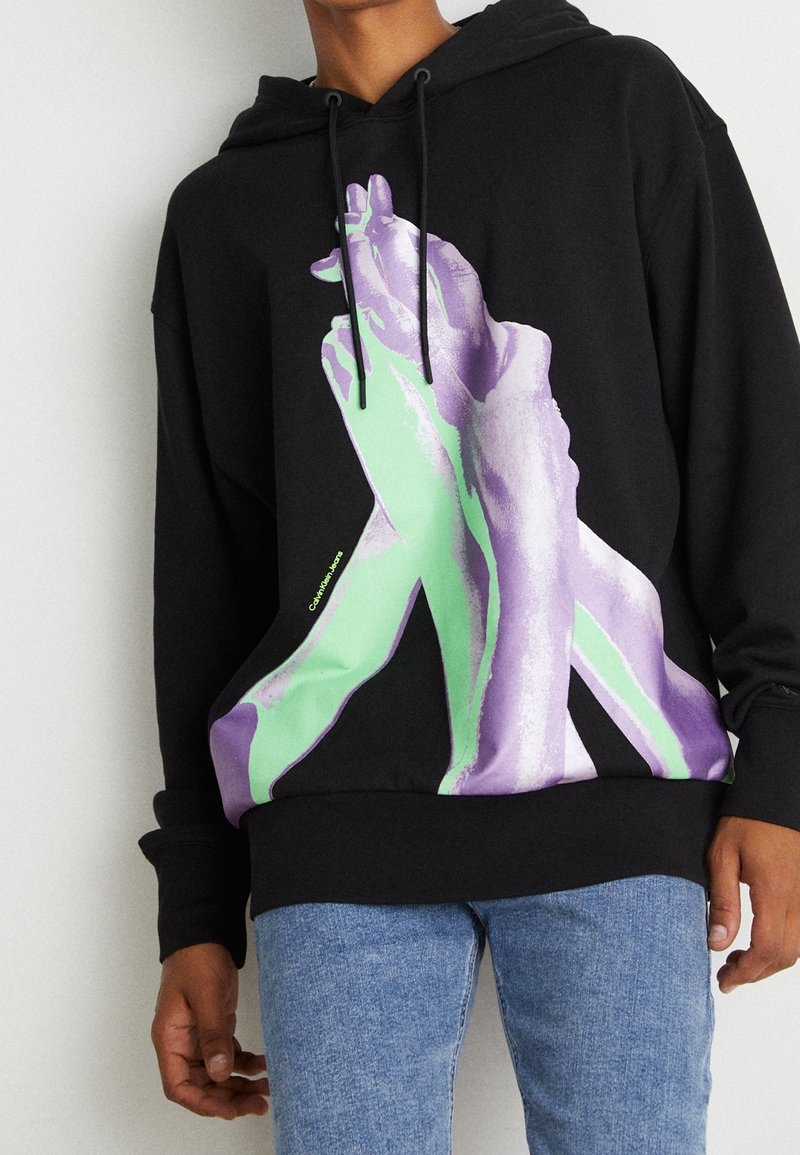 Personne portant un sweat à capuche noir avec un grand graphisme de mains vertes et violettes entrelacées, associé à un jean bleu.