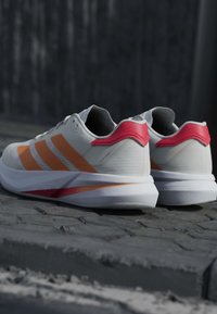 Chaussures de course blanches avec des bandes orange et des touches rouges au talon, placées à l'extérieur sur une surface pavée.