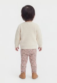 Kleinkind steht mit dem Rücken zur Kamera, trägt einen cremefarbenen Strickpullover, rosa Leggings mit Blumenmuster und beige Knöchelstiefel, vor einem weißen Hintergrund.