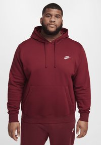 Bordeaux Nike hoodie met een voorzak, een capuchon met trekkoord en een wit logo op de borst. Glad textuur en aansluitende mouwen.