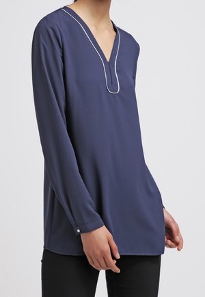 Granatowa tunika z długim rękawem z białym obszyciem wzdłuż dekoltu w kształcie litery V, noszona z czarnymi spodniami na osobie na jasnym tle.