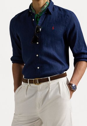 Marineblauwe button-down shirt in lichtgewicht stof, met opgerolde mouwen, gecombineerd met witte geplooide broek en een bruine riem; groene en gemusterde sjaal als accent.