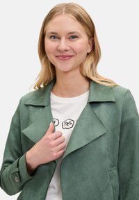 Veste en daim verte avec des revers larges et un design épuré. Ajustée, avec des poignets à bouton, portée sur un t-shirt blanc avec du texte.