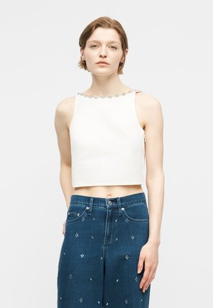 Jeune femme portant un haut court blanc sans manches avec un décolleté décoratif et un jean taille haute bleu orné de petits motifs blancs.