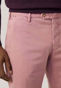 Lichtroze katoenen broek, slim fit, met een knoopsluiting, zijzakken en een gladde textuur. Draagt een donkerpaarse trui.