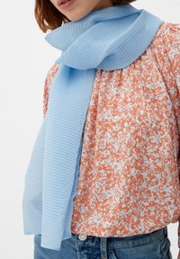 Hellblauer, plissierter Schal aus semi-transparenter Stoff, locker drapiert. Getragen über einem floral gemusterten Hemd in orange und weißen Farbtönen.