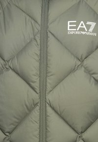 Maslinasto zeleni pufasti prsluk sa preklopom, s prednjim zatvaračem, koji ima suptilni sjaj i EA7 Emporio Armani logo u bijeloj boji na gornjoj lijevoj strani.