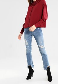 Blusa bordeaux con collo arricciato e chiusura frontale a bottoni, abbinata a jeans chiari strappati e stivali neri alla caviglia con punta affusolata.