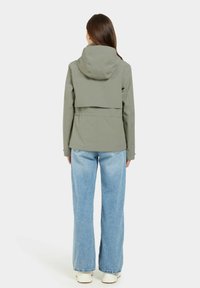 Oliven grøn jakke med hætte, der har et cropped bagdesign og justerbar talje, parret med lyseblå vide jeans og hvide sneakers.