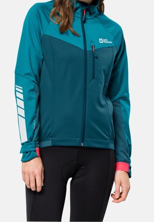 Kvinde iført teal og mørkeblå Jack Wolfskin jakke med lynlås, brystlomme og reflekterende design på venstre ærme, kombineret med sorte cykelbukser.