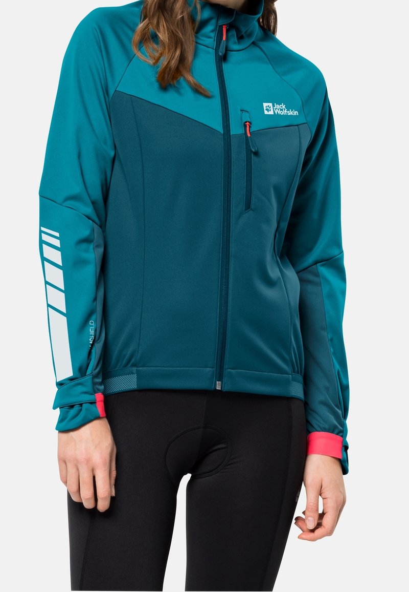 Femme portant une veste zippée Jack Wolfskin bleu sarcelle et bleu foncé avec une poche poitrine et un motif réfléchissant sur la manche gauche, associée à un pantalon de cyclisme noir.