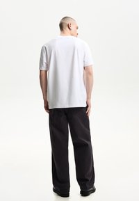 Tricou alb din bumbac, cu mâneci scurte, croială lejeră. Asortat cu blugi negri, cu croială largă și pantofi negri, văzut din spate.