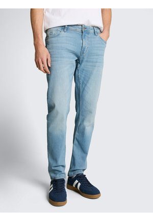 Man draagt lichtblauwe slim-fit jeans en marineblauwe sneakers met witte strepen, staat met één hand in de zak.