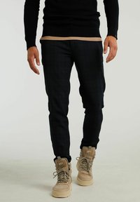 Svarta mönstrade byxor i smal passform som bärs med beige, robusta högstövelkängor. Tröjan i stickad material är mörk och har en texturerad yta.