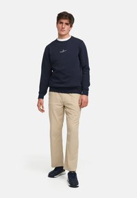 Marineblå sweatshirt med en liten hvit logo på brystet, kombinert med beige bukser og marineblå sneakers. En enkel, uformell design med ribbede mansjetter.