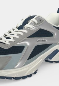 Baskets sportives avec lacets blancs, panneaux en mesh bleu et gris, surcouches argentées et logo Calvin Klein sur le côté.