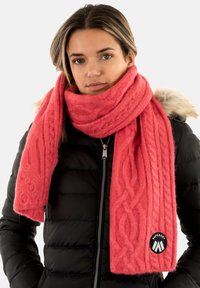 Bufanda de punto coral con patrón de trenzas, hecha de un tejido suave y texturizado, que presenta un parche redondo negro con logo. Combinada con una chaqueta de abrigo negra.