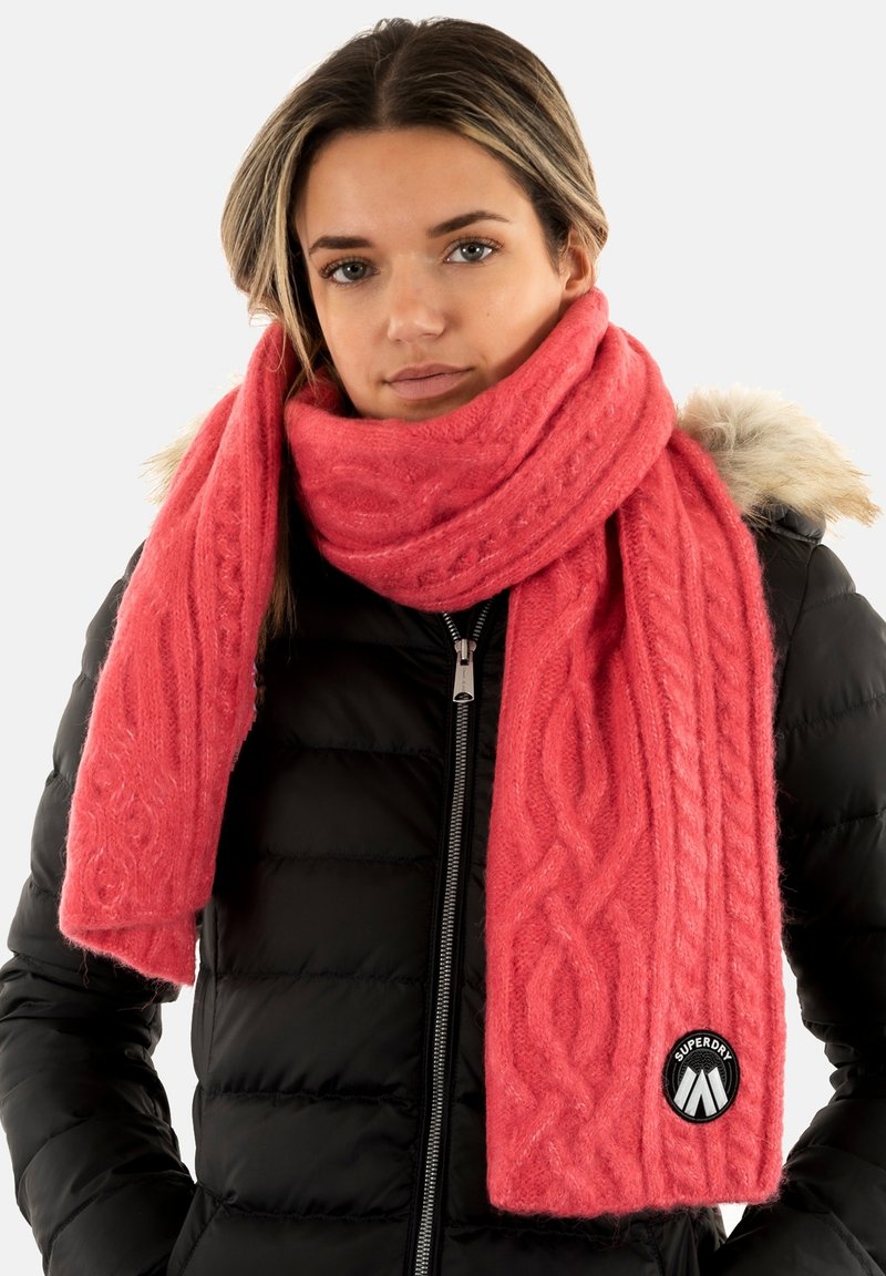 Bufanda de punto coral con patrón de trenzas, hecha de un tejido suave y texturizado, que presenta un parche redondo negro con logo. Combinada con una chaqueta de abrigo negra.