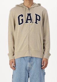 Hoodie beige à fermeture éclair avec lettrage "GAP" en bleu marine, poches avant et capuche avec cordon. Tissu doux et manches longues avec poignets côtelés.