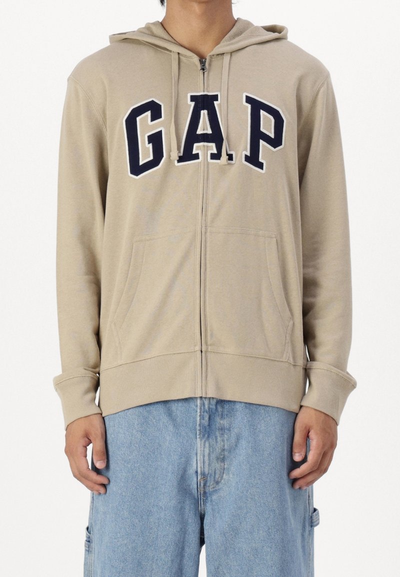 Hoodie beige à fermeture éclair avec lettrage "GAP" en bleu marine, poches avant et capuche avec cordon. Tissu doux et manches longues avec poignets côtelés.