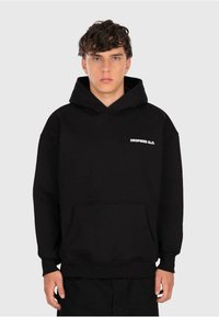 Junger Mann mit lockigem Haar, der einen schwarzen Hoodie mit dem kleinen weißen Schriftzug "DROPSIZE CLO." auf der Brust trägt, vor einem schlichten hellen Hintergrund.