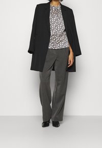 Czarny długi płaszcz, biała i brązowa bluzka we wzory, szare szerokie spodnie i czarne buty tworzą warstwowy, profesjonalny strój.